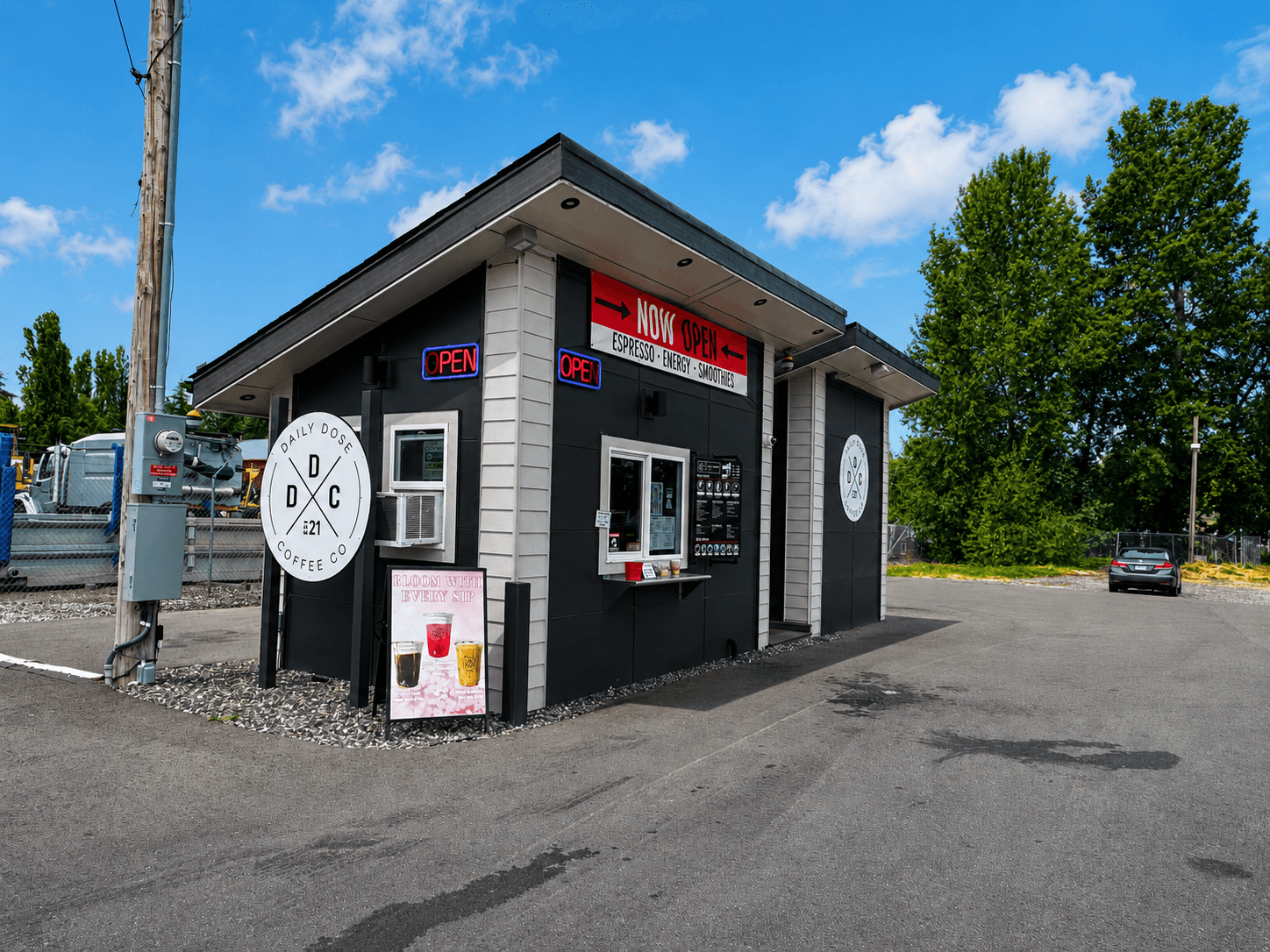A small black and grey drive-thru coffee stand with a neon "OPEN" sign and a "Daily Dose Coffee Co." logo, parked on an asphalt lot.