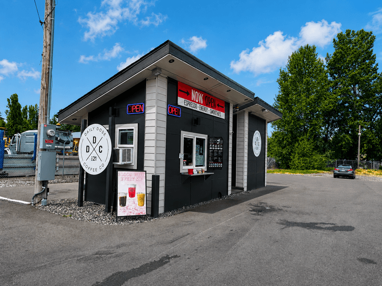 A small black and grey drive-thru coffee stand with a neon "OPEN" sign and a "Daily Dose Coffee Co." logo, parked on an asphalt lot.