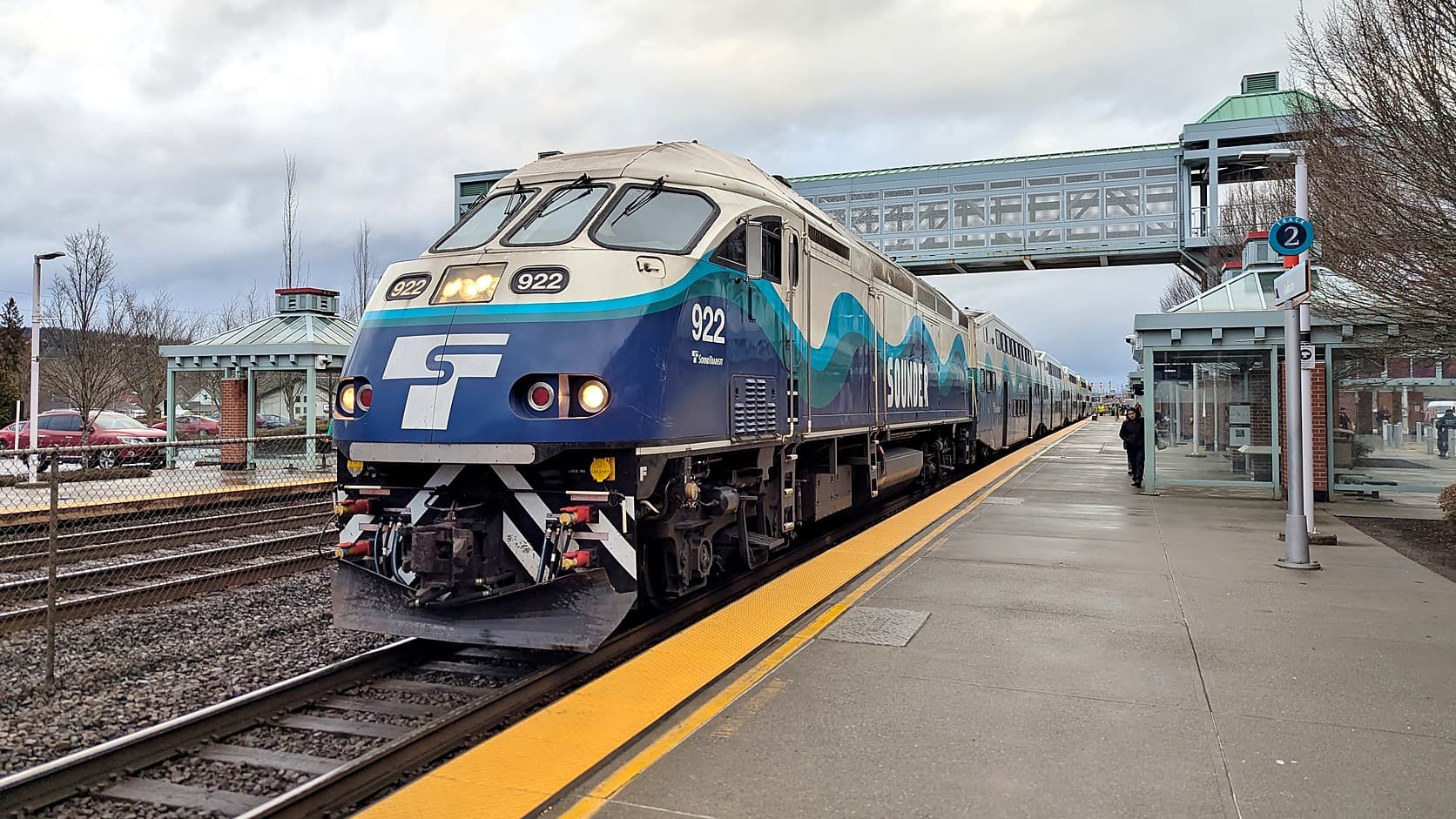 A blue and white Sounder commuter train led by locomotive 922 stands at the platform at Auburn Station under a gray sky, with an enclosed pedestrian bridge crossing the tracks behind it.