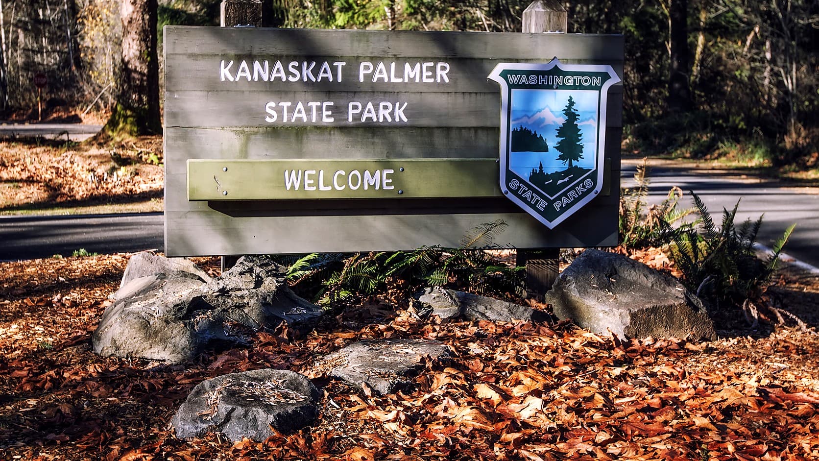 A weathered wooden sign reading Kanaskat Palmer State Park and Welcome, with the Washington State Parks shield, stands behind boulders and fallen leaves at the edge of a forest road.