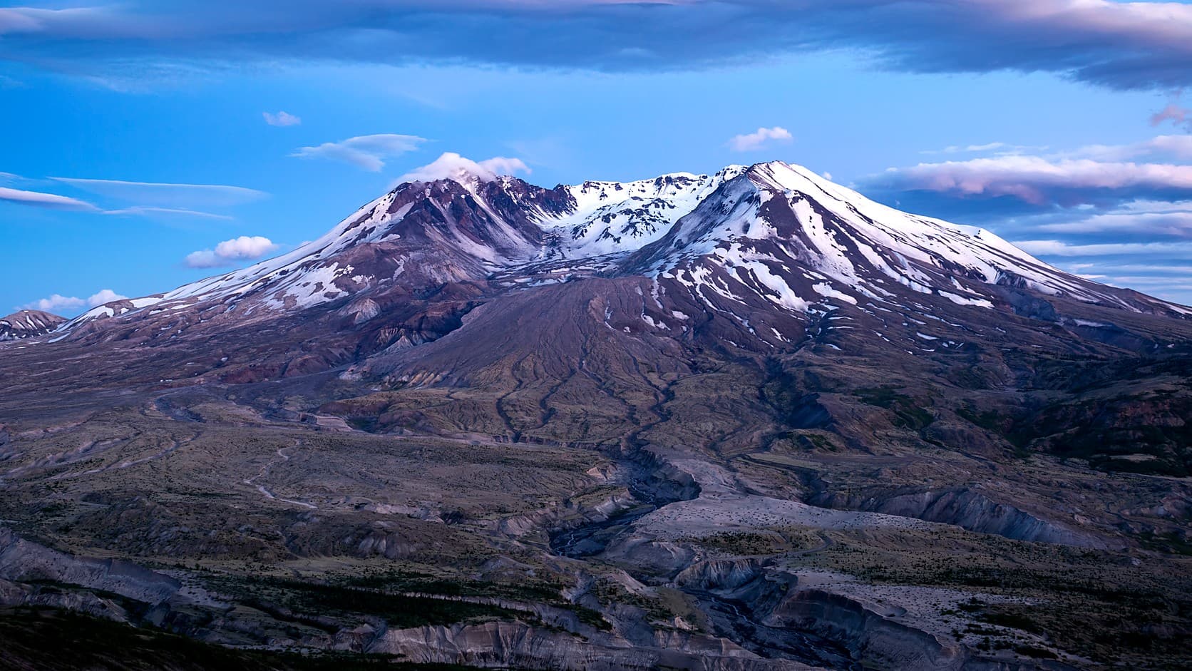 Mount St. Helens at sundown, its snow-streaked summit broken open into a wide crater facing the camera, above gray, gullied ground cut by a braided stream.