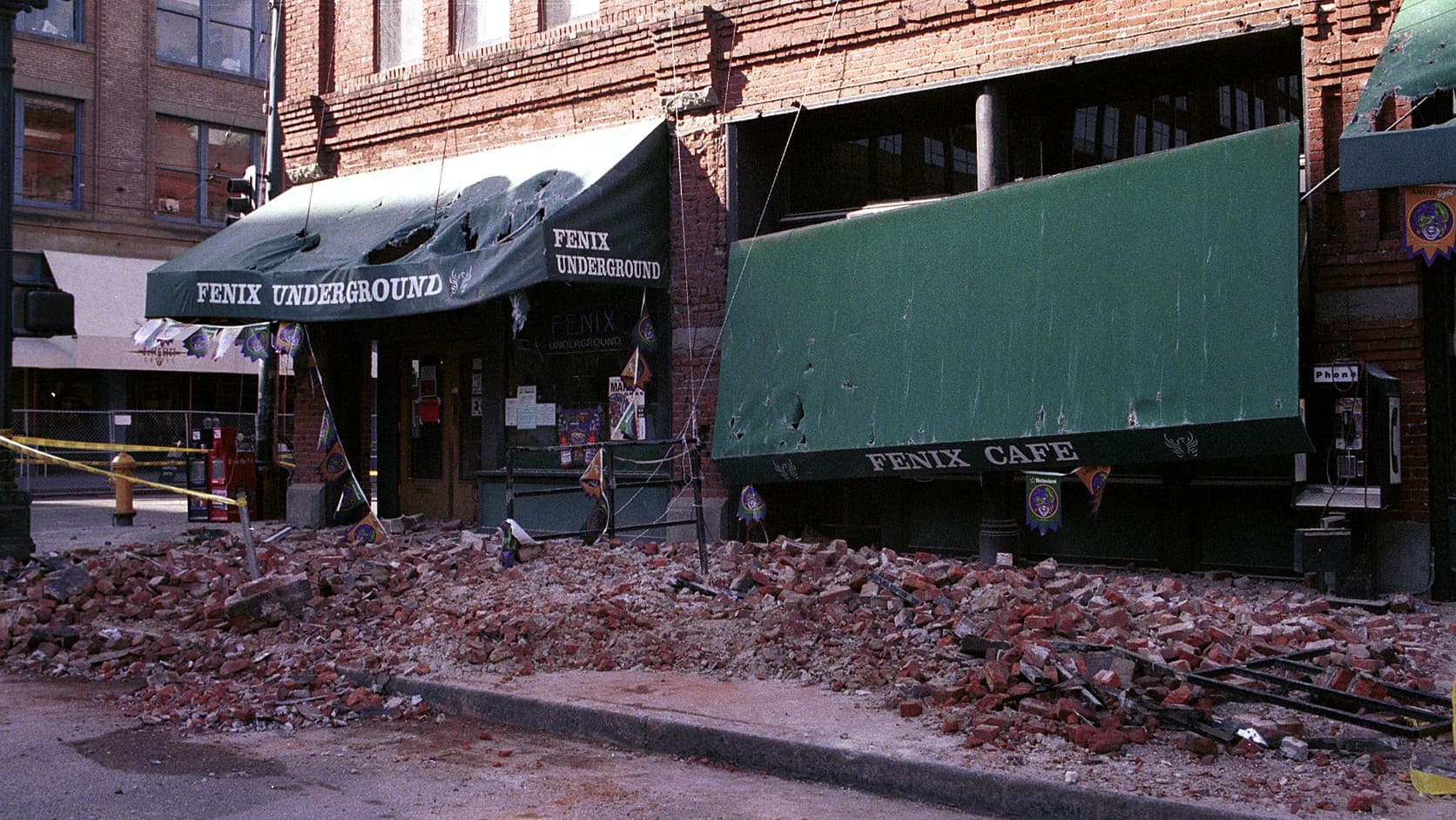 Broken red brick lies heaped across a Pioneer Square sidewalk in Seattle beneath the punctured green awnings of the Fenix Underground and Fenix Cafe, with yellow caution tape strung at left.