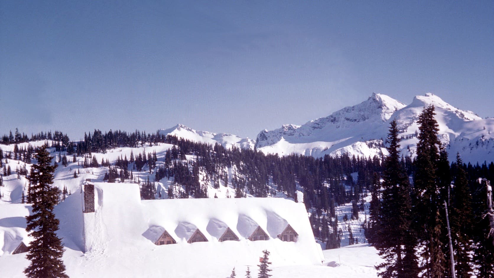 The Paradise Inn at Mount Rainier buried in snow to its dormer windows, with the Tatoosh Range behind it under a clear sky.
