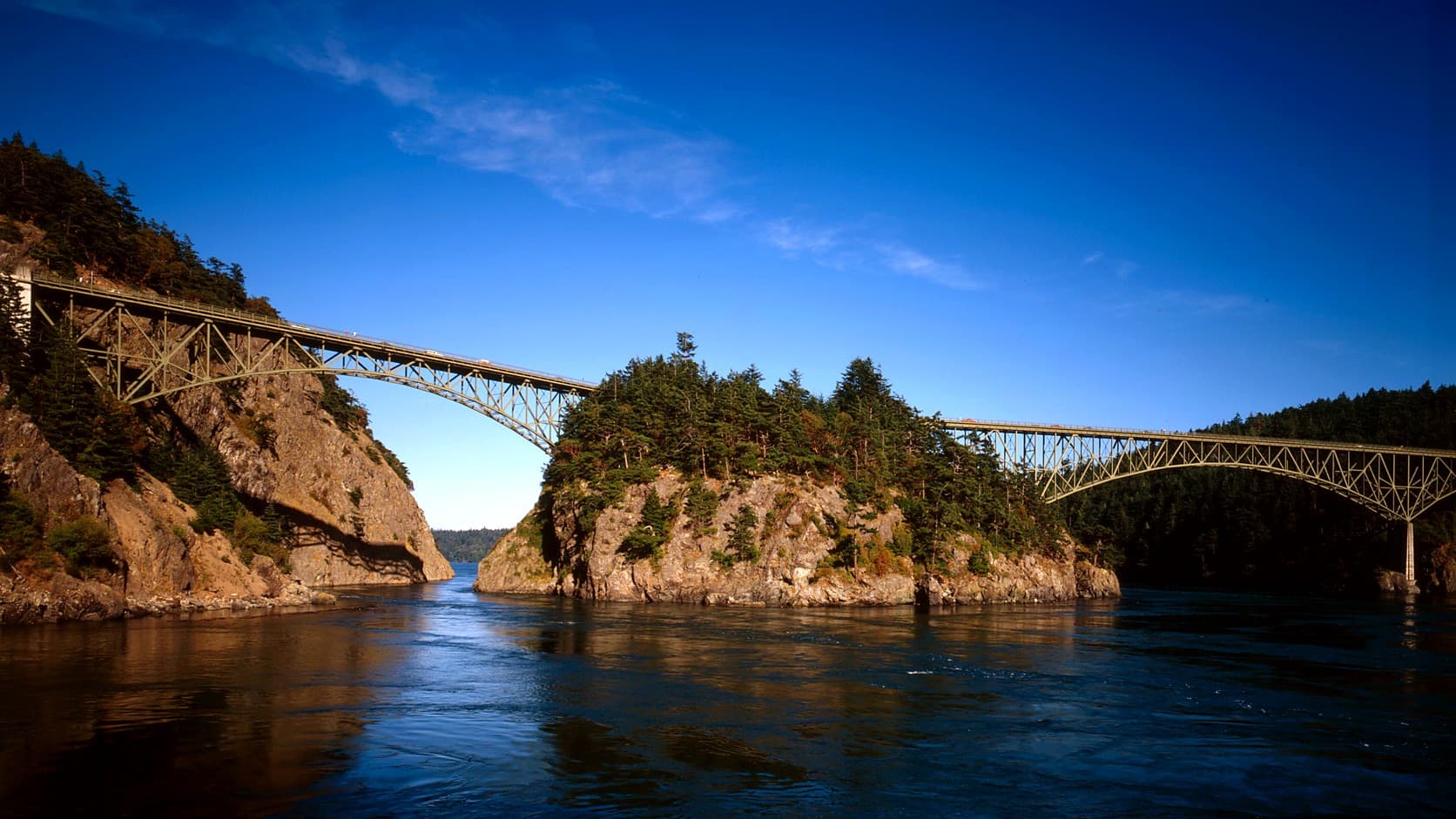 Two steel bridges cross a narrow channel, one running from a rocky, tree-topped cliff at left to a small rocky island crowned with firs and the other continuing from the island to the right edge of the frame, above dark rippled water under a deep blue sky.