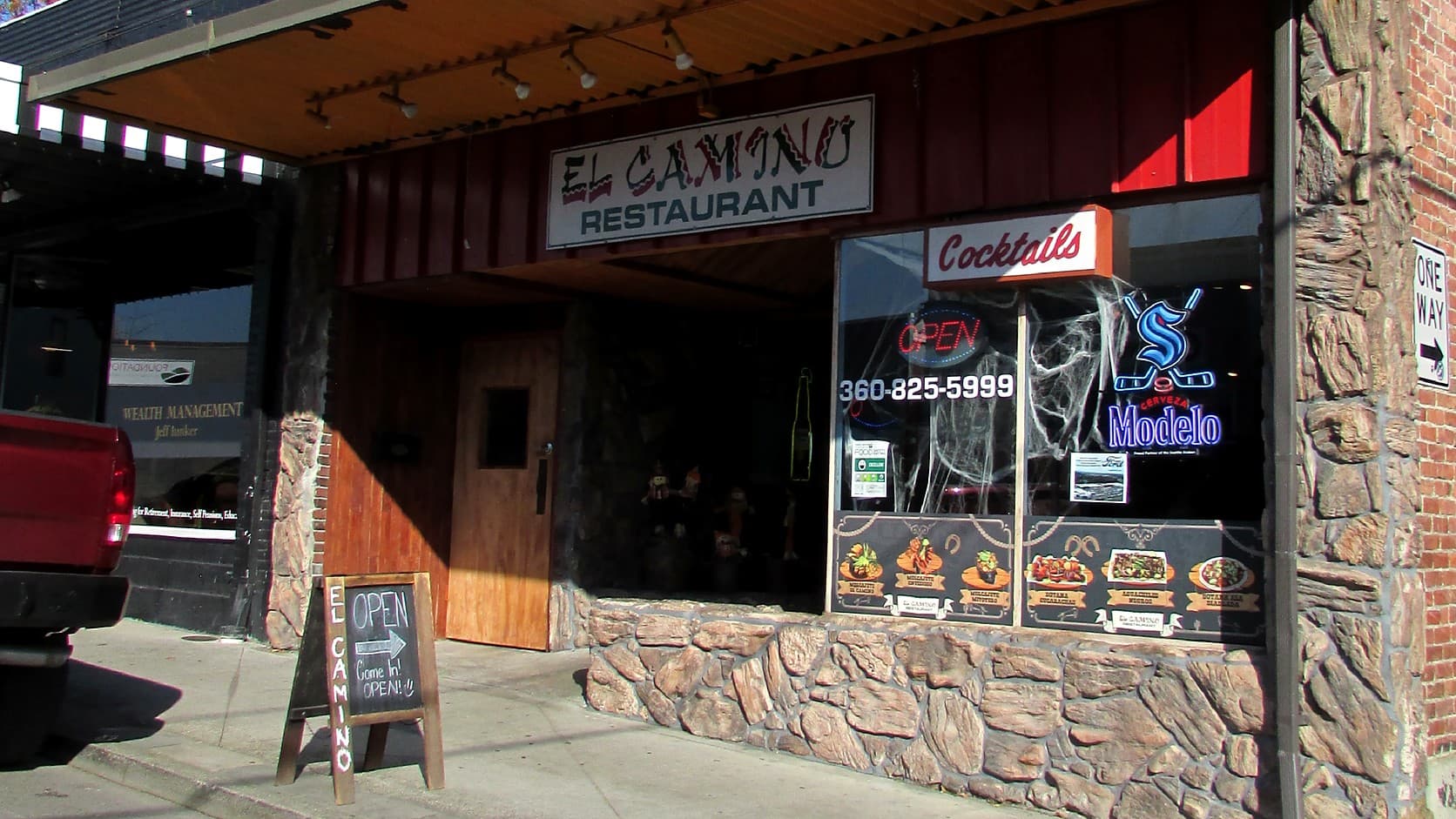 The front of El Camino Restaurant in sunshine: its sign under a metal awning, a wooden door, and a window with a Cocktails sign, an Open sign and the phone number 360-825-5999 above food posters, with a chalkboard reading Open on the sidewalk.