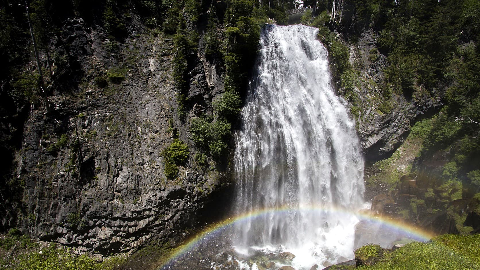 Narada Falls pours white water down a dark rock cliff in Mount Rainier National Park, with a stone bridge at its top and a rainbow arching through the spray at its base.