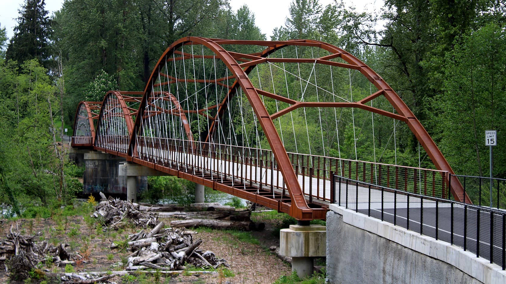 The three rust-red steel arches of the Foothills Trail bridge over the White River at Buckley, Washington, seen from one approach in May 2025, with driftwood on the riverbank below.