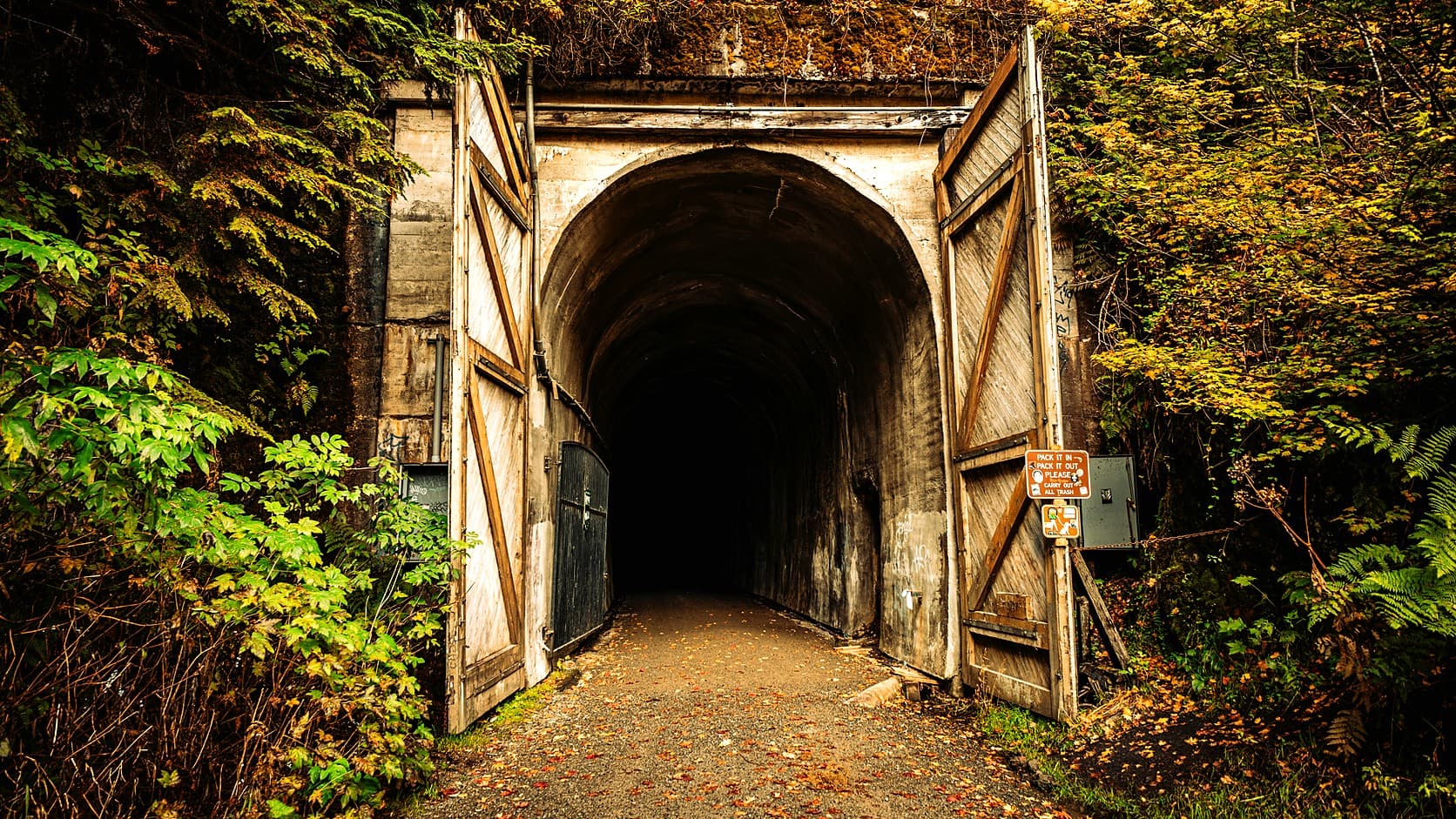 The arched concrete east portal of the Snoqualmie Tunnel at Hyak, with its wooden doors open and a gravel trail scattered with fallen leaves running into the dark, framed by autumn foliage.