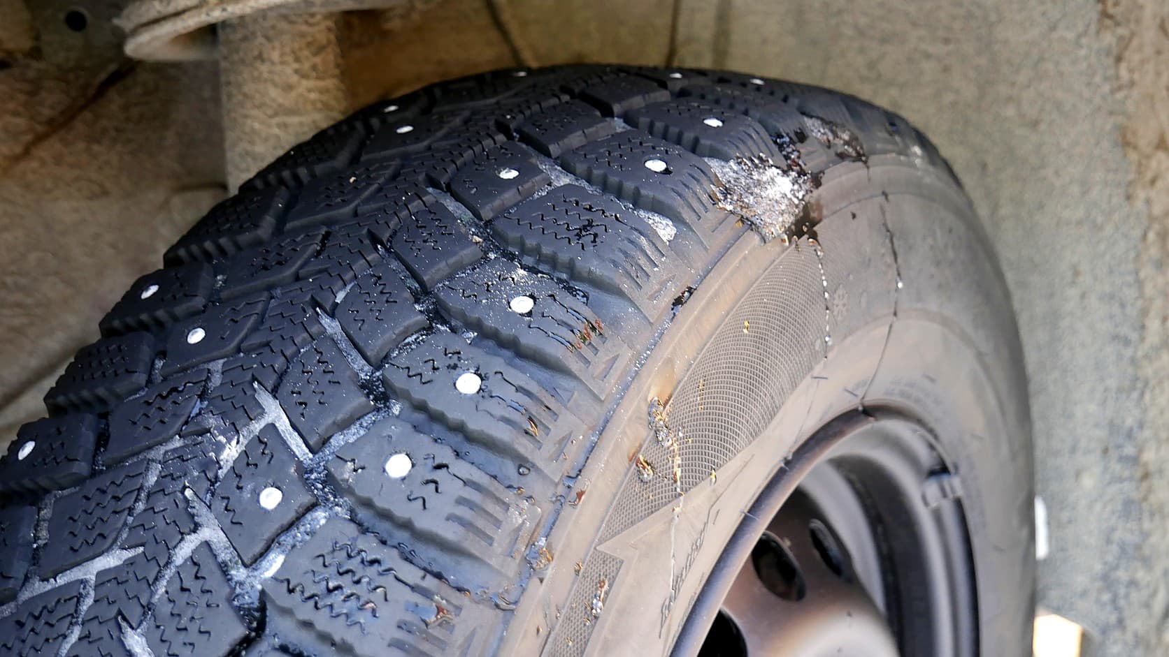 A winter tire with rows of small metal studs set into its tread blocks, seen in a car's wheel well beneath a dusty coil spring.