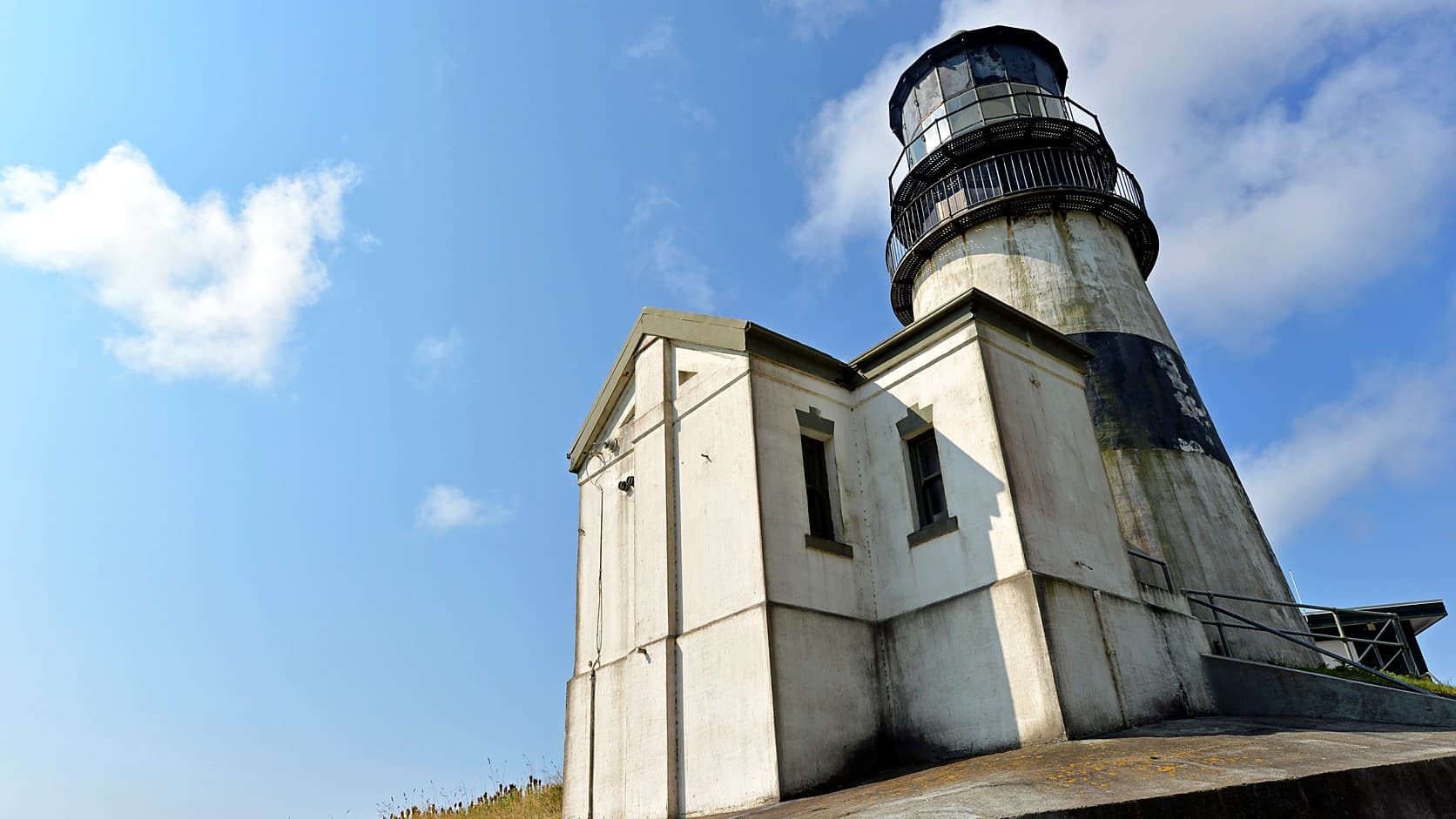 The Cape Disappointment Lighthouse seen from below, a white tower with a black band and a glass lantern room rising behind a squat attached white outbuilding under a blue sky.