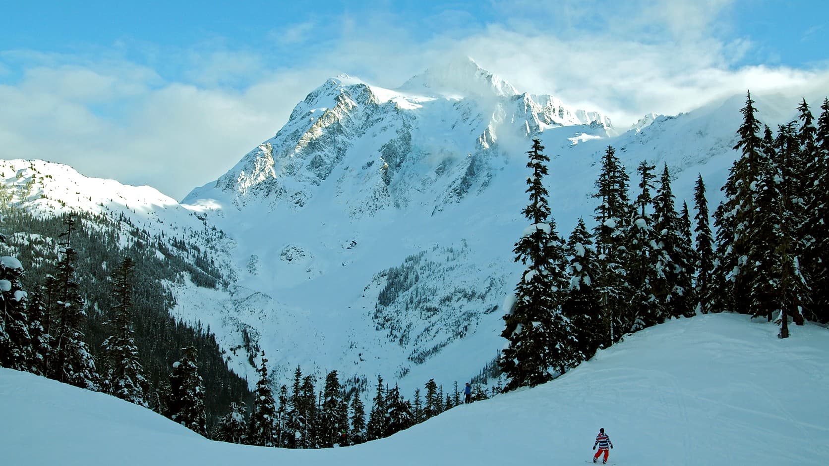 A snowboarder on a snow-covered slope far below Mount Shuksan, whose snowy summit rises above dark evergreens under a blue sky with thin cloud.