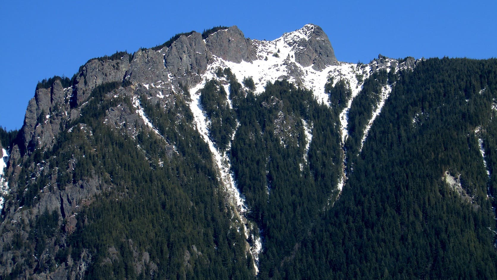 The rocky, snow-streaked summit ridge of Mount Si rises above dark fir forest under a clear blue sky.