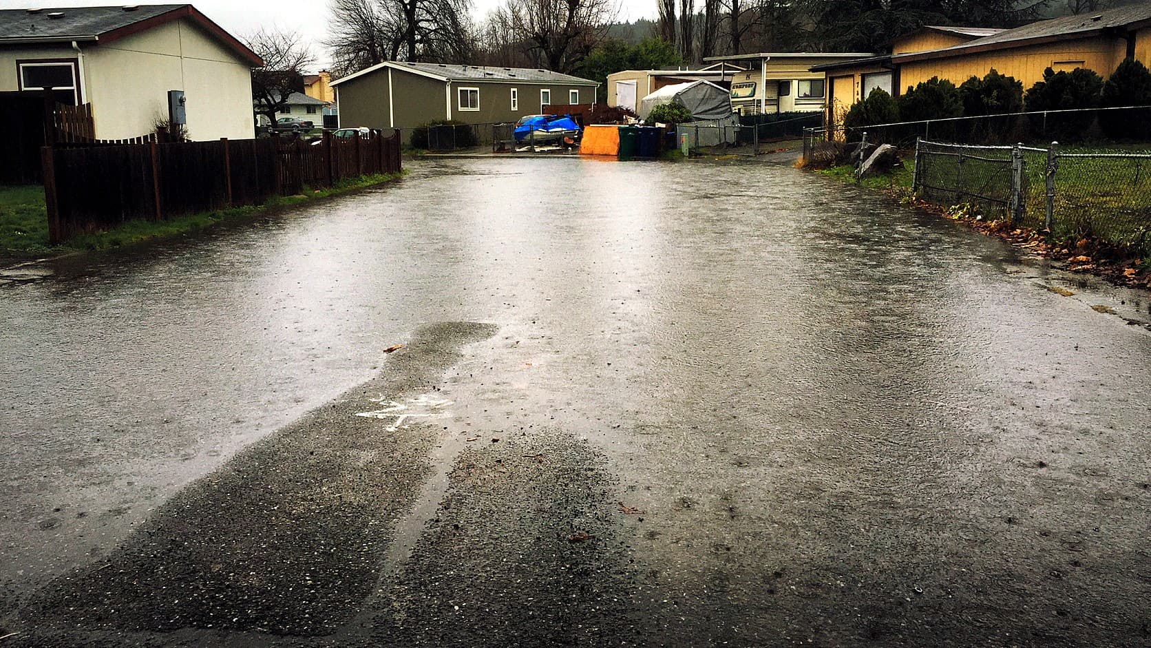 A rain-soaked residential street in Pacific, Washington, with water sheeting across the asphalt, wooden and chain-link fences and small houses on both sides, and bare trees behind.