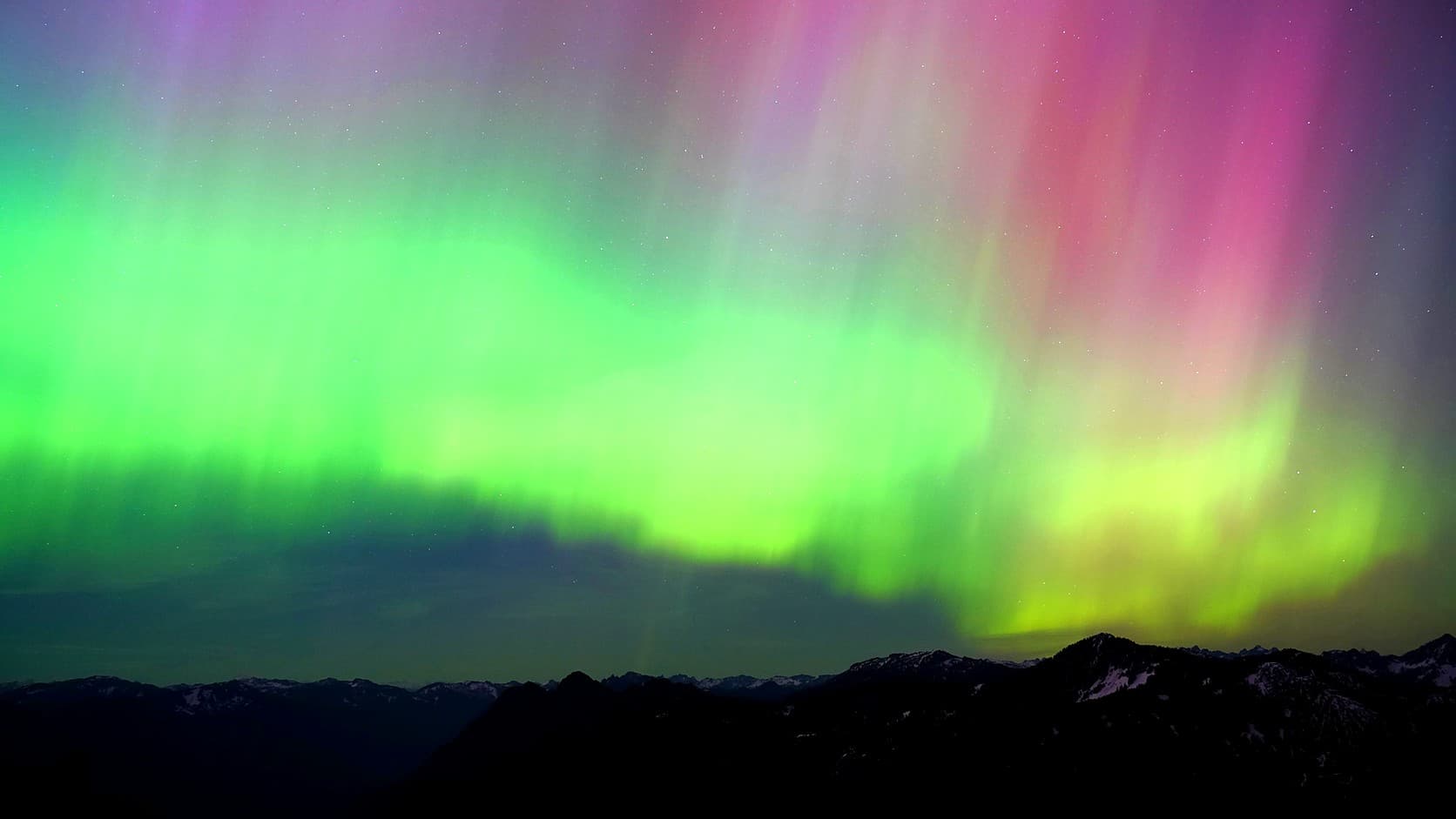 A broad band of green aurora, with pink and violet rays rising above it, arcs across a starry sky over a dark mountain ridgeline with patches of snow.