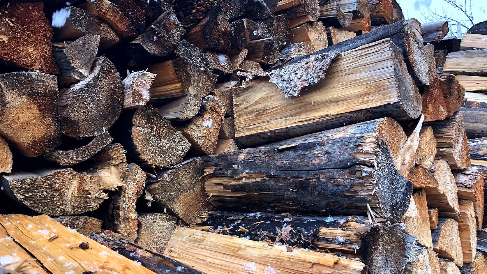 A chest-high wall of split and round firewood with bark and sawn ends toward the camera, snowflakes in the air and a gray sky above.
