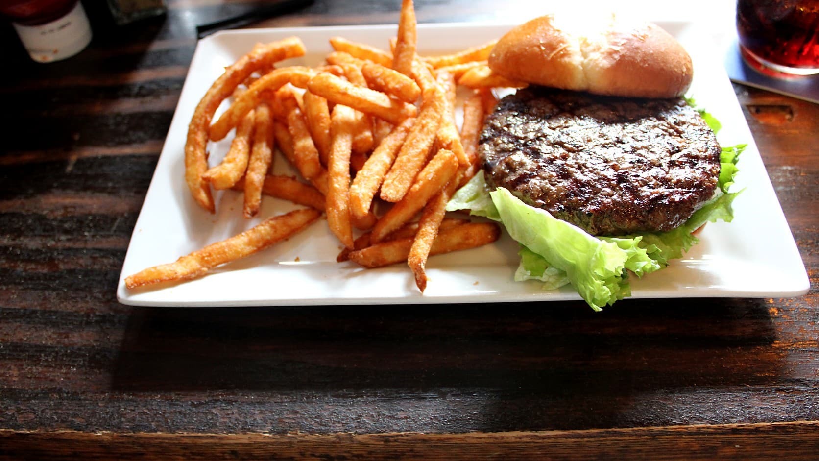 A grilled burger patty on green leaf lettuce, with the top of its bun set behind it, beside a heap of fries on a white square plate on a dark wooden table.