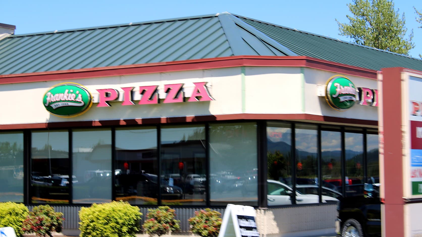 A single-story pizza restaurant with a green metal roof, red trim and a cream front bearing green oval Frankie's signs and red PIZZA letters, its plate-glass windows above a row of shrubs.