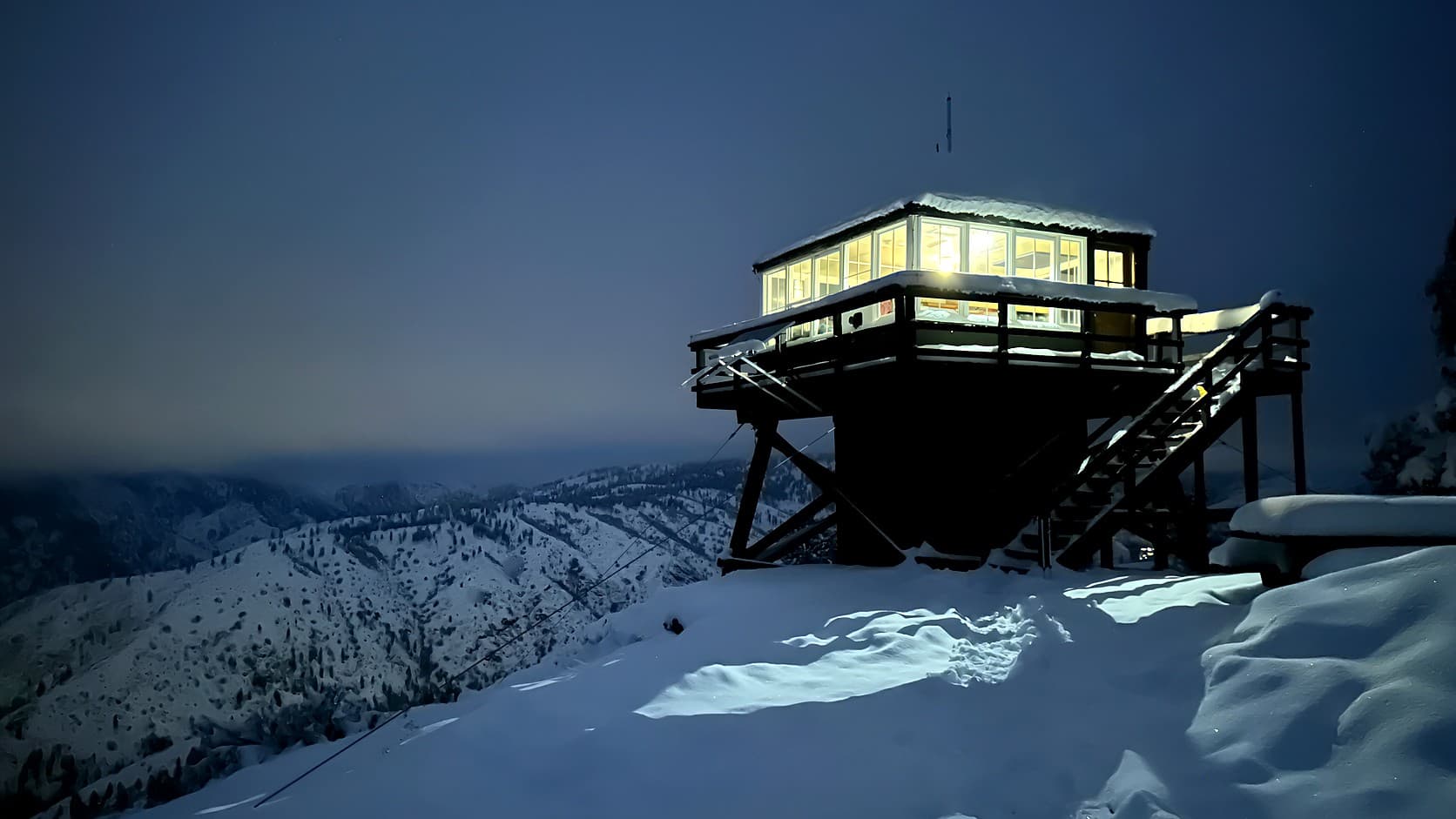 A small lookout cab with windows on every side glows from inside atop a short timber tower, its railed deck and outside stairs dusted with snow, above deep snow and snowy, tree-dotted ridges under a dark blue night sky.