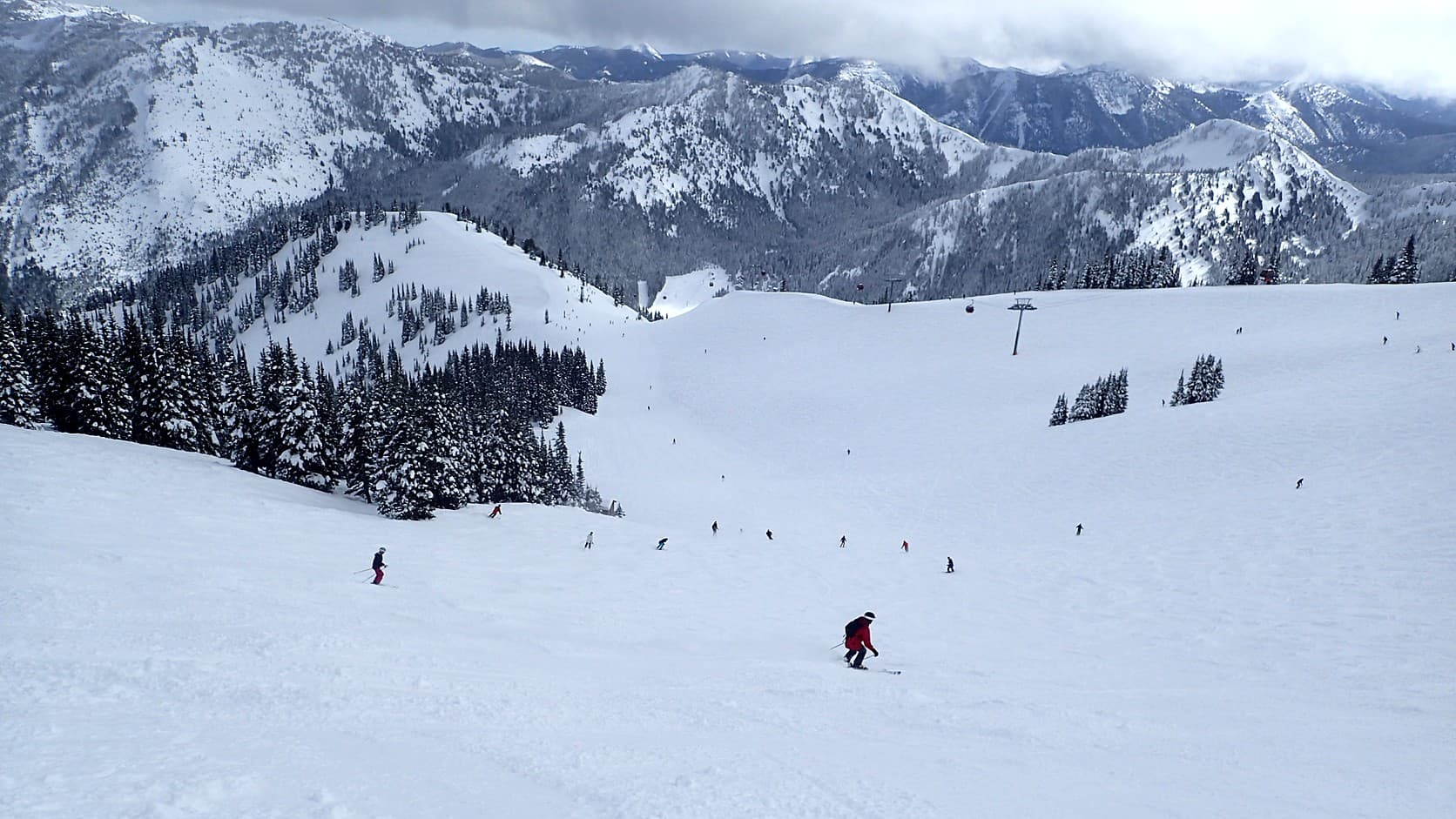 Skiers spread down a wide groomed run on Crystal Mountain under low cloud, with red gondola cabins and a lift tower in the middle distance and snow-covered ridges beyond.
