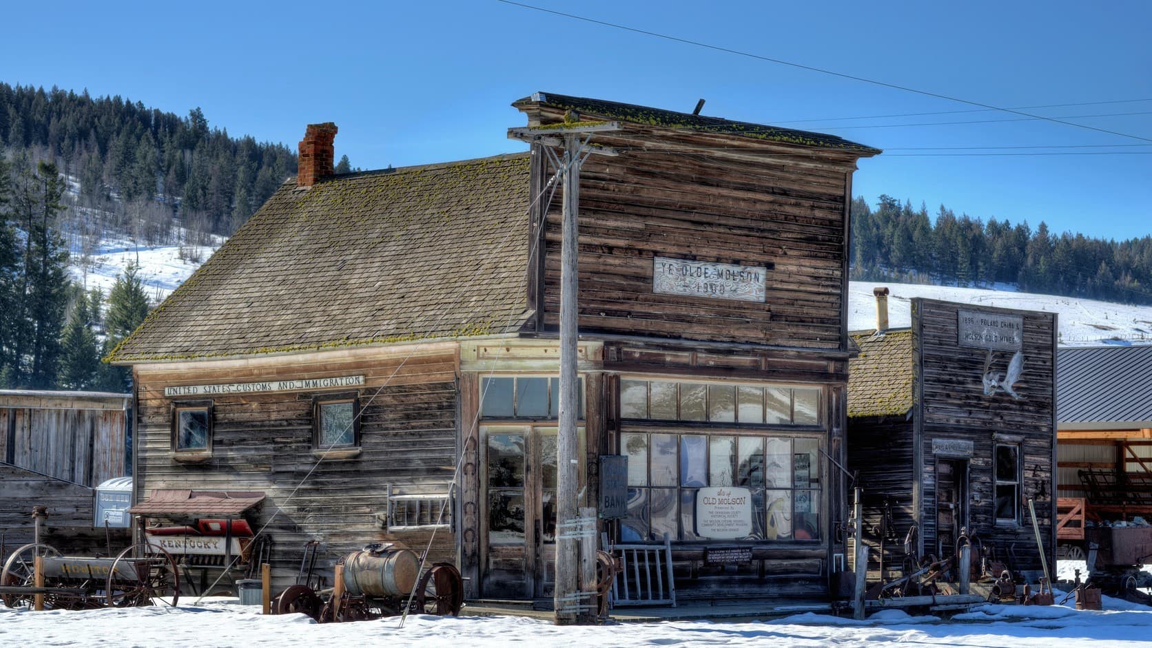 A weathered false-front wooden bank building with a sign reading Ye Olde Molson 1900, joined to a gabled wing signed United States Customs and Immigration, with old wagons and machinery standing in snow under a clear blue sky.